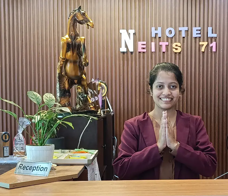 A smiling receptionist greets guests with folded hands at the Hotel Neits front desk